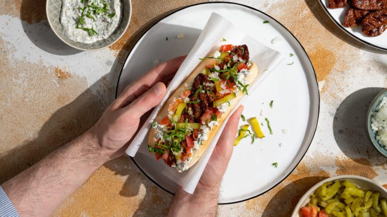 Sucuk Hot Dog wird in den Händen gehalten, bereit zum Essen. Daneben stehen Schälchen mit Toppings.