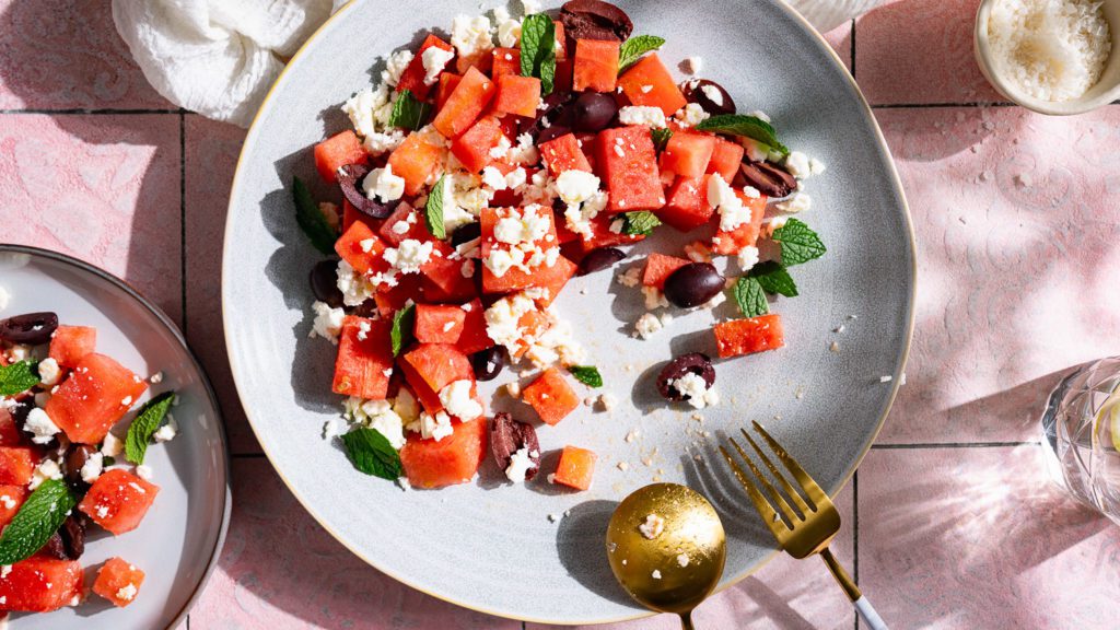 Melonensalat mit Feta, Minze und Oliven auf einem großen Teller angerichtet. Der Untergrund ist rosé farben.