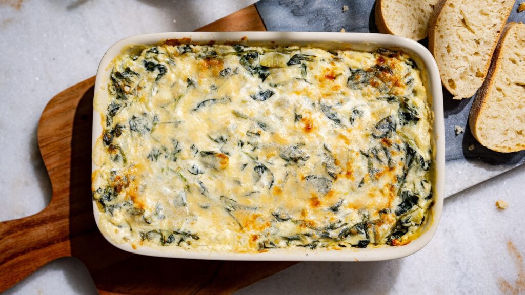 Spinach and artichoke dip in a casserole dish. Next to it is a sliced baguette.