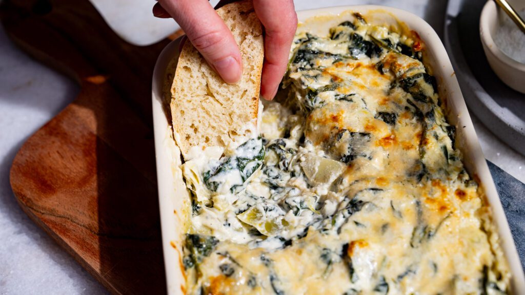 One hand is dipping a piece of baguette into a casserole dish with spinach and artichoke dip.  