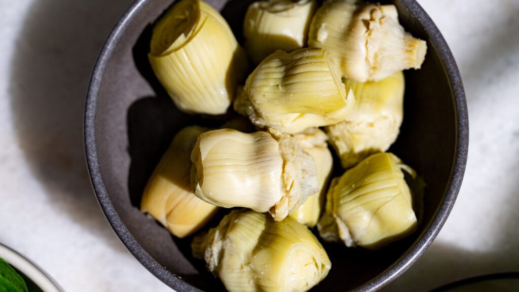 Artichoke hearts in a dark bowl on a light marble table.