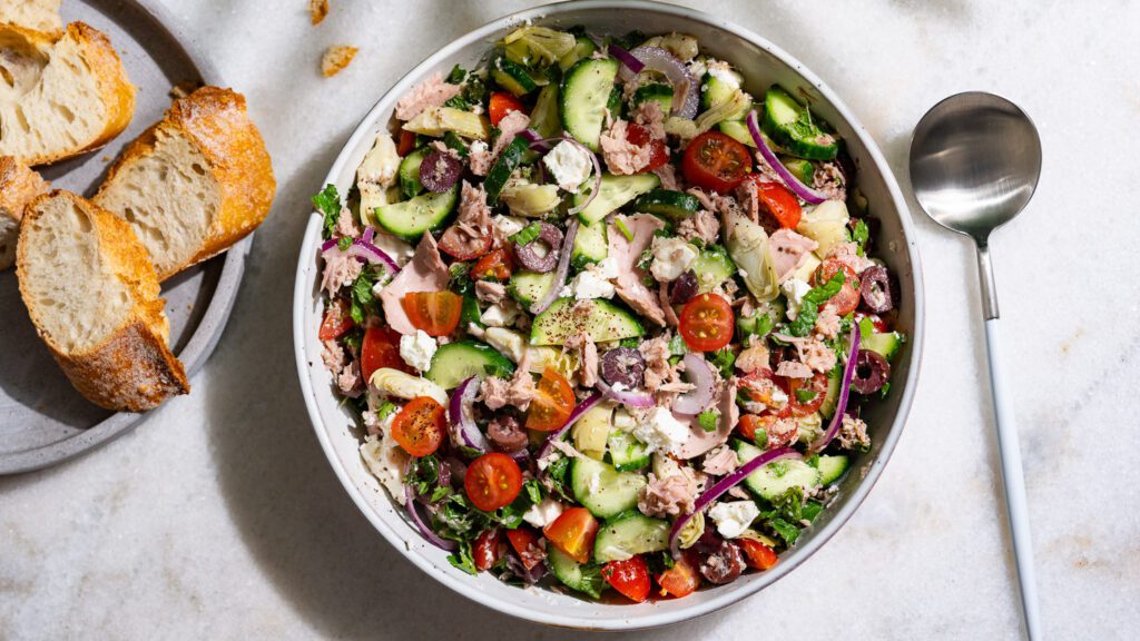 Ensalada mediterránea de atún servida en un cuenco. Hay una baguette en un plato al lado.