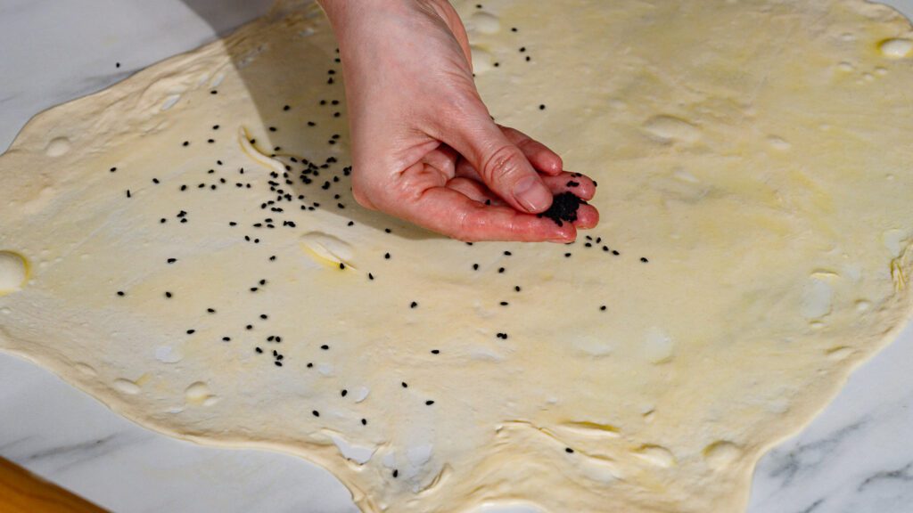 Dünn ausgerollter Malawachteig wird mit Butter bepinselt und mit Schwarzkümmelsamen bestreut.
